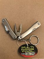 Photo of silver metal multi purpose pocket tool with pliers open, a small LED light built into one arm, and a keychain attached, placed on a cardboard background.