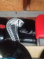 Black Shelby Cobra seat back showing the white Cobra snake logo and Carroll Shelby signature on the surface.