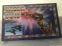 Front view of the original box featuring the branding 'Laser Gun', description of battery operated with 4 laser sounds and blinking lights, and artwork of a person holding the toy gun.