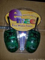 Photo shows two green translucent plastic Emac walkie talkies with black antennas in original packaging backing. The packaging displays the Emac logo and product name, advertising over 200 feet range.