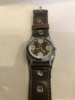 Close-up photo of the watch face showing dial, numerals, metallic bezel, leather strap with metal grommets.