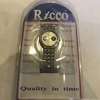 Photo showing Ricco brand wristwatch in original packaging with clear plastic cover. Dial has white background with two distinct black and white semicircles and Ricco logo. Stainless steel linked band.
