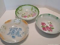 Three hand painted porcelain bowls with floral designs and scalloped edges arranged in a row showing different color rims and flower patterns