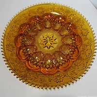 Top-down photo showing the amber glass egg dish with detailed floral and geometric pressed patterns, 12 molded egg sections, and a serrated edge.