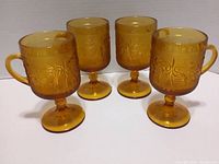 Front view showing four amber glass Tiara Sandwich tall mugs with pedestal foot and embossed design