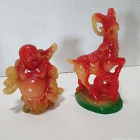 Both resin figurines together showing Feng Shui Coined Goat and Laughing Buddha in red, yellow, and green colors
