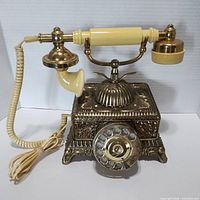 Front view of vintage grand emperor rotary dial telephone showing ornate brass base, rotary dial, cream colored handset and cord.