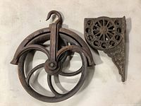 Two antique metal pulleys showing the larger one marked No 12 with hook and curved spoke wheel and the smaller pulley with decorative bracket