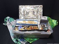 Overall view of plastic container holding assorted costume jewelry including necklaces, pins, bracelets, and a small presentation box with pins, as well as the abalone tile jewelry box in background on a decorative scarf.