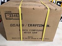 View of the front side of the cardboard box with product identification label showing 10 inch compound miter saw by Sears/Craftsman, with yellow plastic straps securing the box.