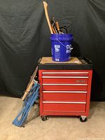 Red Craftsman rolling toolbox with blue bucket of hand tools on top and folded metal Workmate leaning against side