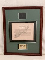 Framed artifact showing the coin in a small window, certificate, and plaque inside wooden frame with green and black mats.