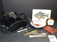 Photo showing the assortment including Swift Triton binoculars with case, fish art on rag paper, Linden travel alarm clock, pens, and a small leather-bound book.