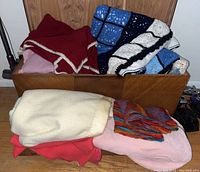 Overall view of blankets including burgundy knit, blue and white crocheted, cream, pink, red wool blankets on and near wooden chest.