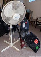Photo showing a white Windmere standing fan next to a burgundy and gray Hoover Power Scrub carpet cleaner with a black mesh bag containing a hose and fittings.