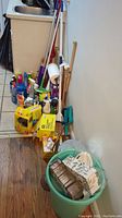 View of cleaning supplies including mops, cleaner bottles, and box packages lined on floor next to wall.