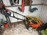 Side view of the red and black electric lawnmower with yellow extension cord on top, placed on a shed floor with other garden tools in the background.