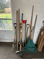 Assortment of garden tools standing upright against a wall, including rakes, spades, hoe, broom, and edger.