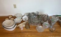 Wide view of all items including Corningware dishes, hand-painted Azoza ceramics, crystal and clear glassware, vases, and assorted serving dishes and stemmed glasses on wooden floor against white wall.