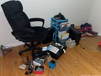 Photo of black office chair with padded armrests on wheeled base, blue plastic drawer organizer filled with papers and office supplies, black HP printer/scanner on floor, multiple office supplies scattered including phones, staplers, and calculators