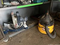 Photo showing the MasterCraft miter saw on a metal shelving unit next to the Stinger wet/dry vacuum on the floor.