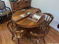 Round oak dining table with two leaves on top and five wooden spindle-back chairs arranged around it in a room with wood flooring.