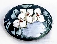Front view of oval porcelain brooch showing hand painted white flowers with green leaves on dark background.