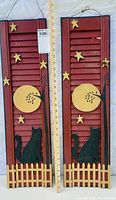 Two tall red wooden shutters side by side, each with hanging wire bracket, black cat silhouette sitting on yellow wooden fence at bottom, large yellow moon and yellow stars affixed on shutter slats