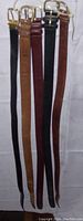 Five men's leather belts hanging vertically, showing various colors and brass buckles.