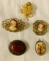 Five vintage brooches and one pendant displayed on white background showing cameo carvings, painted portraits, stone settings, and decorative metal filigree frames.