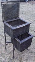 Front view of antique wicker sewing stand showing open lift-up top storage compartment and front pull-out drawer basket, standing on four legs.
