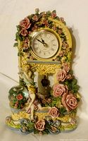 Front angle view of cherub angel floral tabletop clock showing detailed rose decorations, clock face with Roman numerals, cherub figurines and ornamental base.