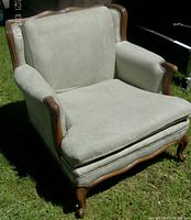 Side angle view of a plush light teal fabric armchair with walnut wood accents on a grassy outdoor ground, showing cushioning and detailed wooden legs and frame.