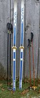 Top side view of white Inovik skis with blue and black logos and gold 3-pin bindings along with two sets of poles.