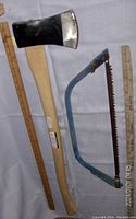 Photo showing the full length of the axe with wooden handle and metal head alongside a ruler and the blue frame bow saw with serrated blade.