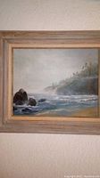 Front view of framed seascape painting showing rocky shore and ocean with trees on cliffs.