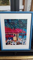 Framed artwork showing colorful outdoor cafe scene with people seated under a red awning and tree canopy