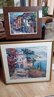 View of two framed paintings stacked on floor showing colorful European village and coastal landscape scenes with frame damage and scratches