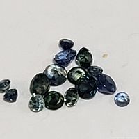 Multiple small faceted blue sapphire gemstones scattered on a white surface, showing different shades and cuts.
