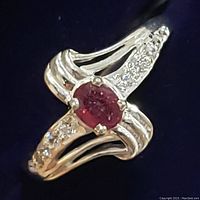 Close-up image of sterling silver ring with oval ruby and clear accent stones on band, on black background.