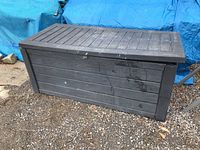 Closed large black plastic deck box with wood plank design on the exterior, placed outdoors on gravel.