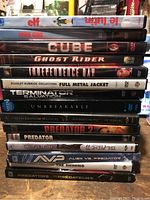 Stack of 15 DVDs showing titles Elf, Young Guns, Cube, Ghost Rider, Independence Day, Full Metal Jacket, Terminator Salvation, Unbreakable, Texas Chainsaw Massacre, Predator 2, Predator, Blade II, Alien vs Predator, The Shining, and Predators.