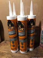 Five orange and black Gorilla Construction Adhesive tubes with pointed plastic nozzles standing upright showing full tube at front with Gorilla logo and product details visible.