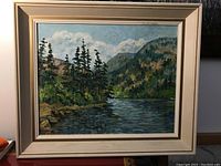 Framed landscape painting showing river, rocks, trees, and hills under sky.