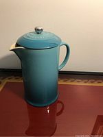 Front view of the blue ceramic Le Creuset French press on a reflective surface.