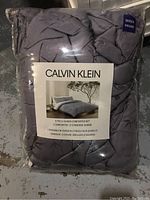 Photo of packaged Calvin Klein queen comforter set showing quilted gray comforter and two shams inside original plastic packaging with brand label.