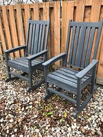 Two dark grey resin rocking chairs side by side outdoors on gravel against a wooden fence