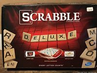 Front of the box showing 'Scrabble Deluxe' branding, a depiction of the rotating gameboard, and mentioning carry case and non-slip grid features.