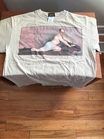 Full view of beige Seinfeld graphic t-shirt with large front image of George Costanza in a comedic seductive pose on a wooden table.
