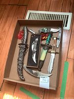 Box containing multiple decorative and ornate knives, some with sheaths; includes a curved blade with a green handle, a silver ornamental sheath knife, and others.